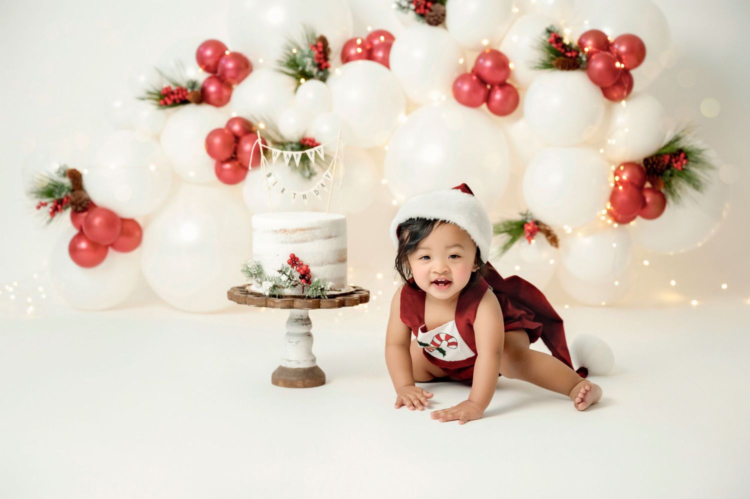 Baby in Santa hat crawls near Christmas cake with white and red balloon backdrop and festive holiday decorations.