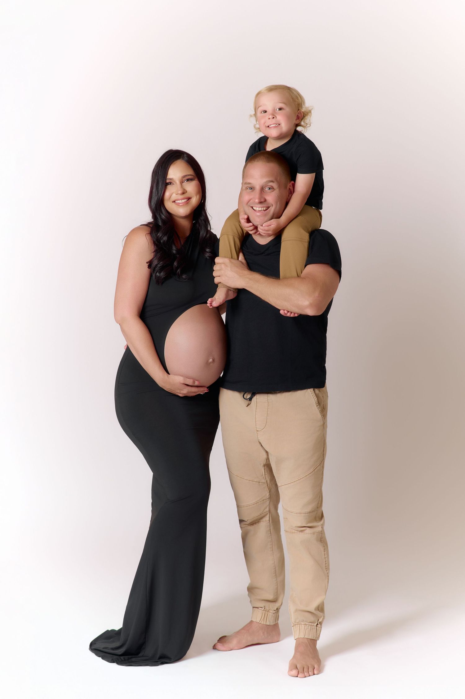 A pregnant woman in a black gown poses with her partner and young child on his shoulders in a studio maternity photo.