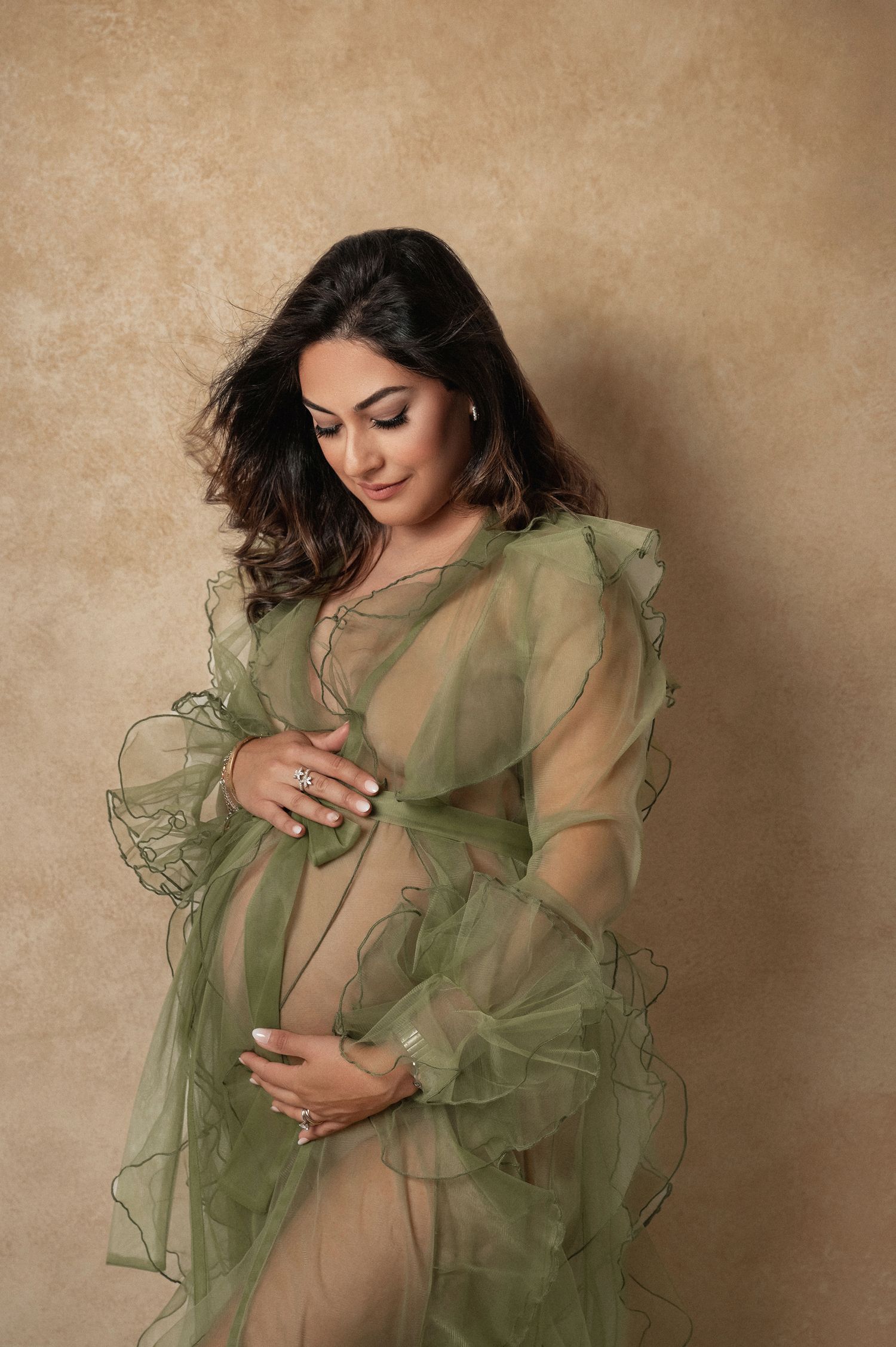 Pregnant woman in sheer green ruffled gown cradling baby bump, elegant maternity portrait against warm beige background.
