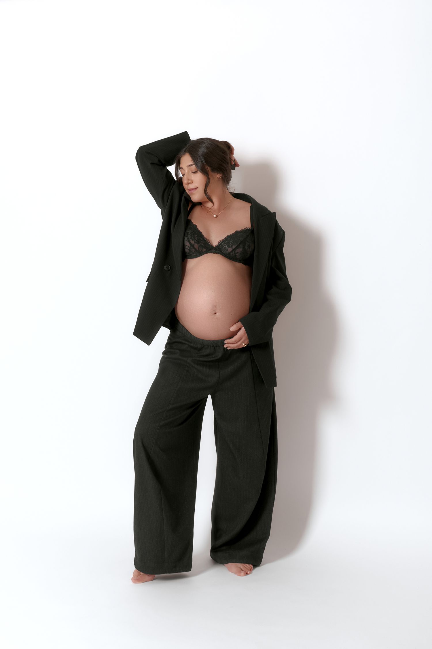 Pregnant woman in black blazer and wide-leg pants with lace bra in studio maternity shoot.