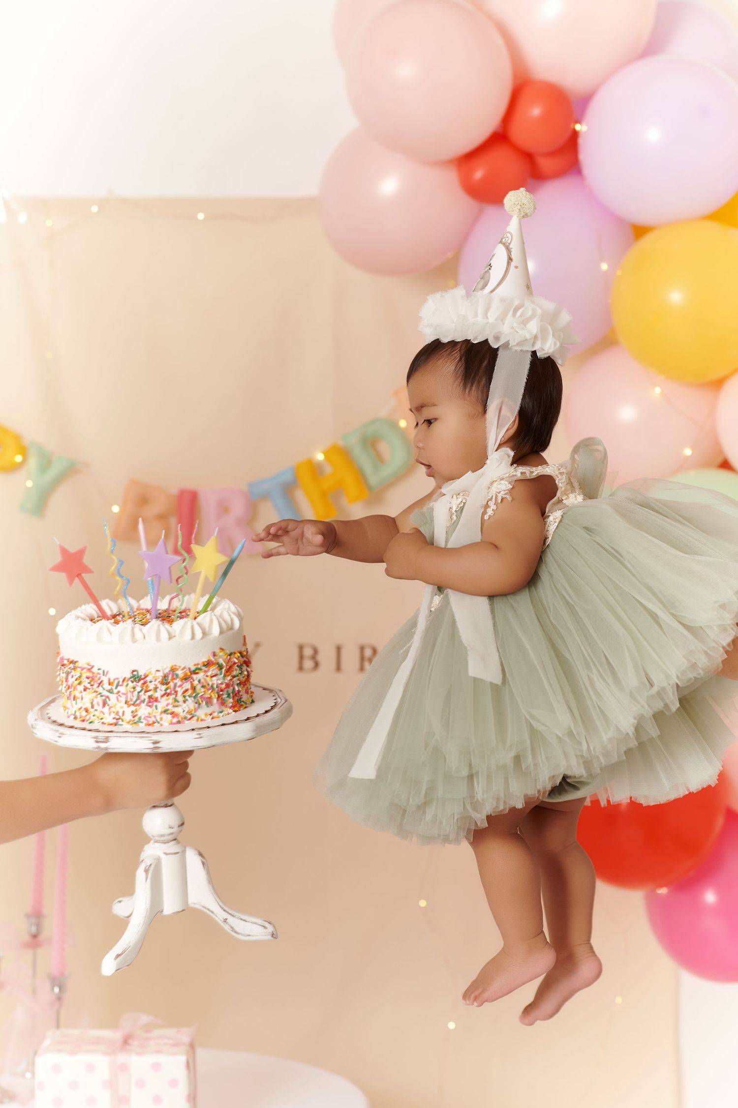 Baby girl in green tutu dress reaching for birthday cake with colorful sprinkles, surrounded by pastel balloons.