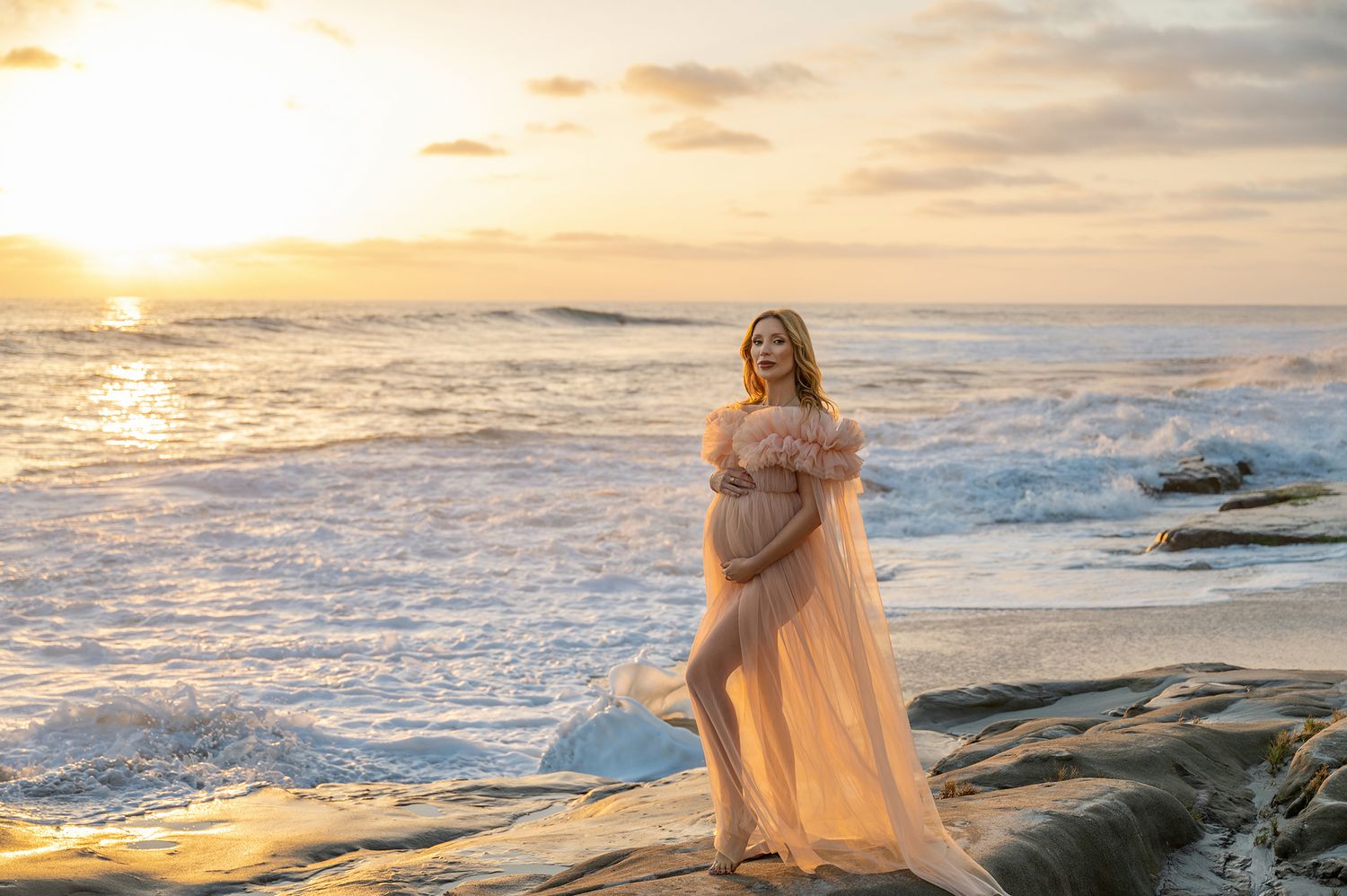 Pregnant woman in blush tulle gown standing on rocky shore at golden sunset by the ocean.