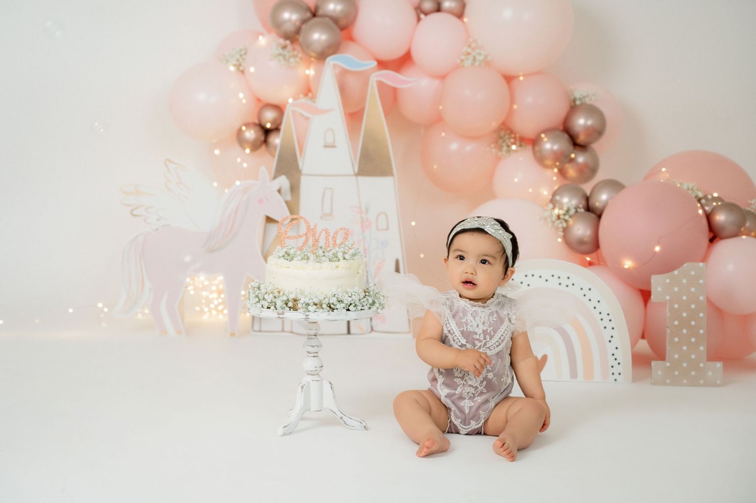 Baby girl first birthday cake smash photo with pink balloons, castle decor, and glitter outfit in studio setting.