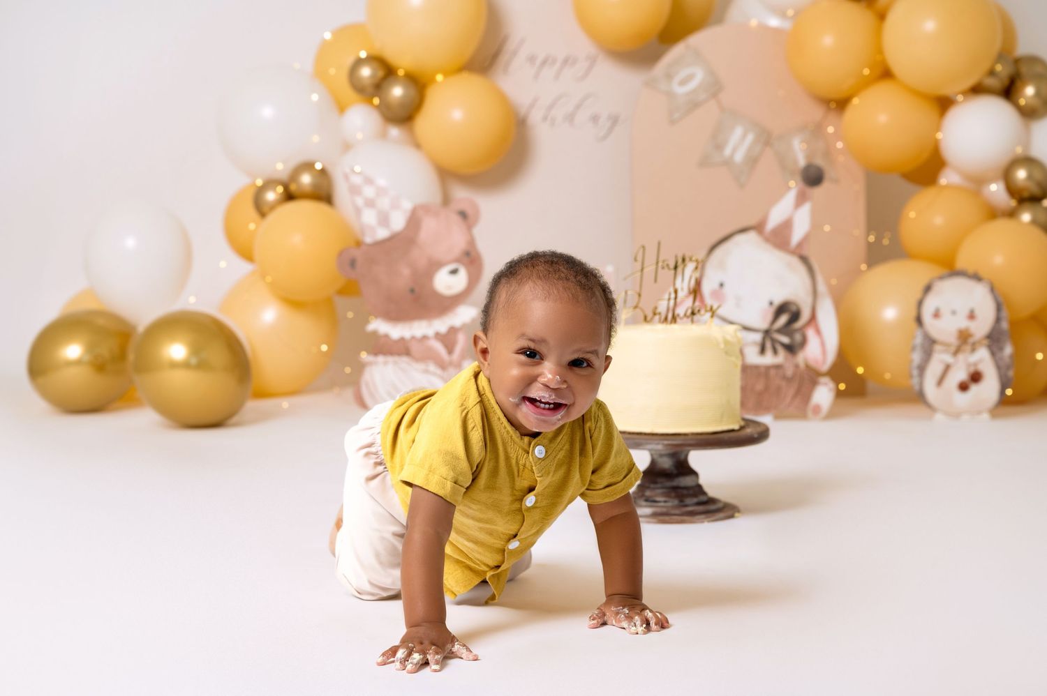 Baby boy in yellow outfit crawls toward first birthday cake with gold balloon arch backdrop.