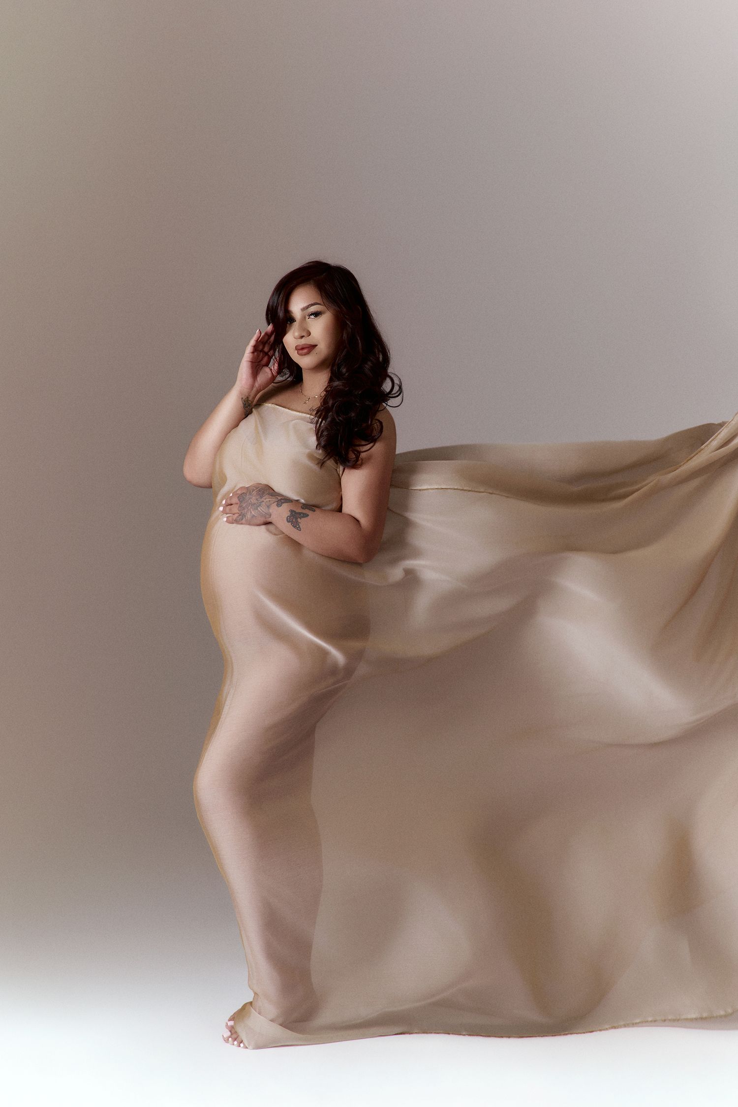 Pregnant woman draped in flowing beige silk fabric, showcasing baby bump in an artistic maternity San Diego studio portrait.