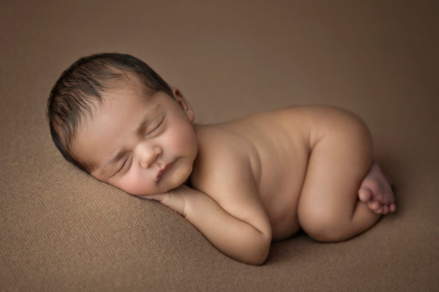 Peaceful newborn baby sleeping curled up on a soft brown blanket in a classic posed newborn photography style.