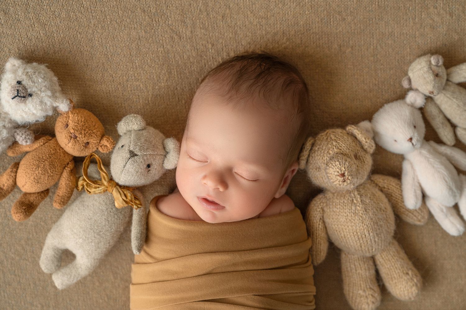 Sleeping newborn baby wrapped in mustard yellow swaddle surrounded by soft teddy bears on beige background.