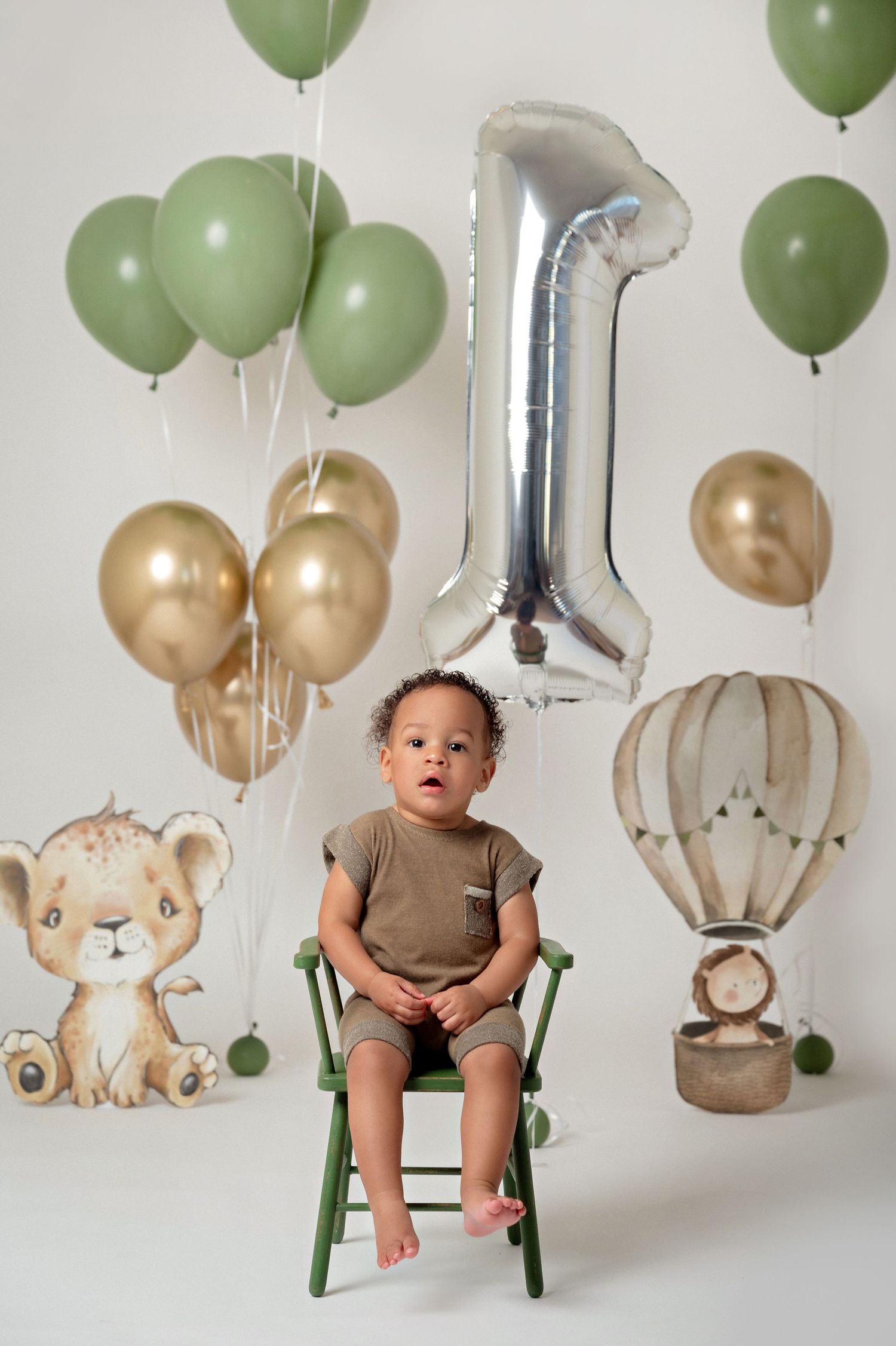 A baby sits on a green chair surrounded by sage green and gold balloons with a silver number 1 balloon for a first birthday.