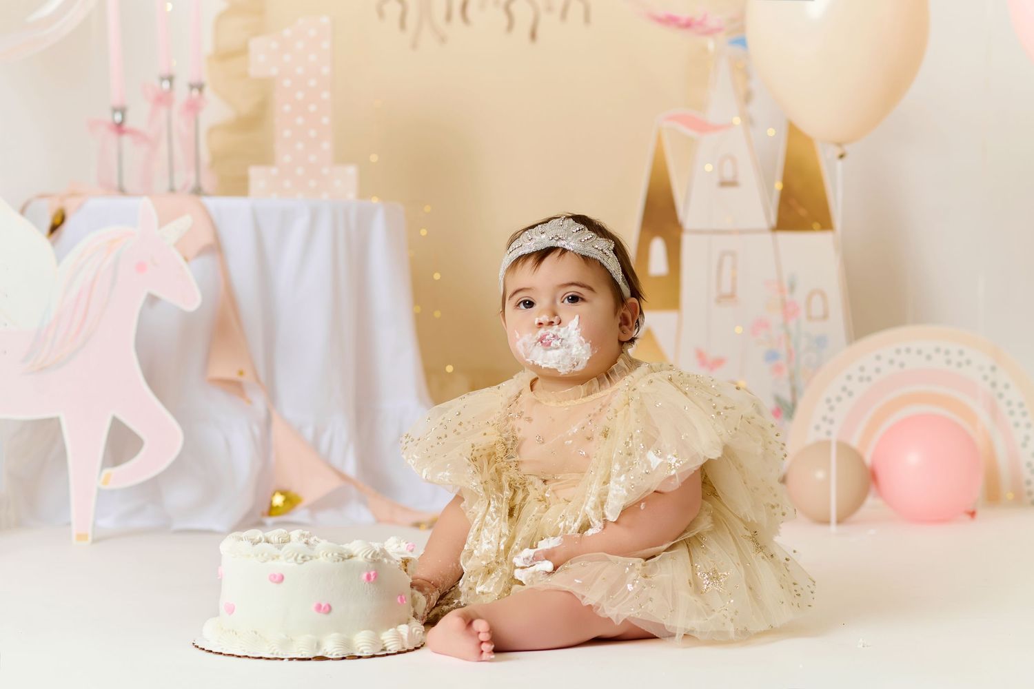 Baby girl in gold dress with cake on her face celebrating her first birthday with unicorn decorations.