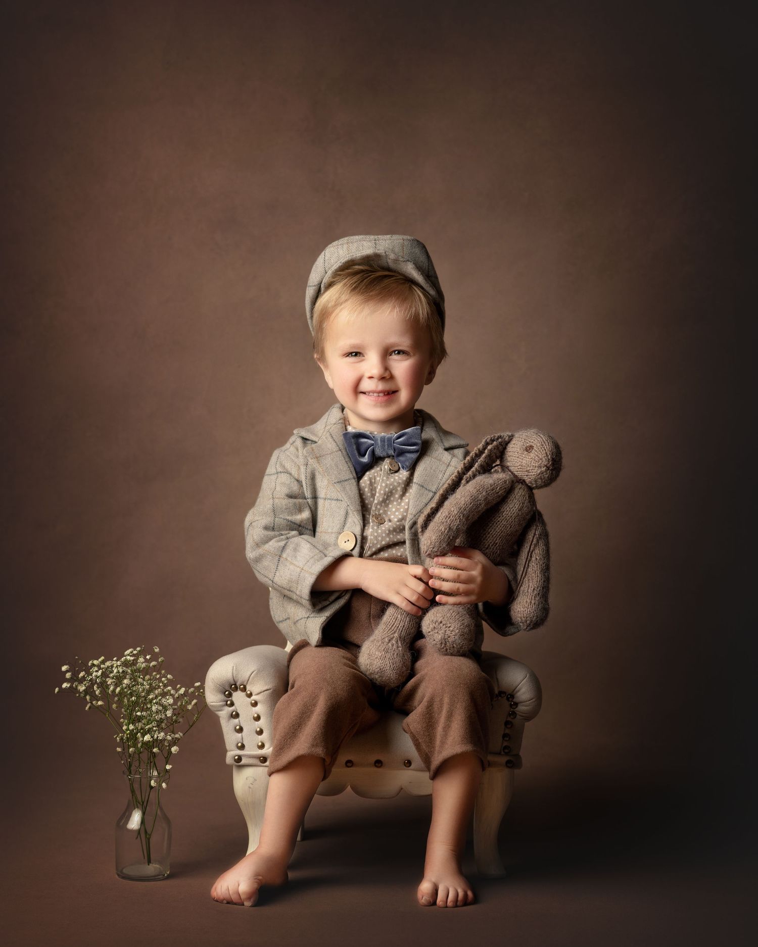 Smiling toddler boy in gray suit, bow tie, and cap holds stuffed bunny while seated on vintage chair with flowers nearby.