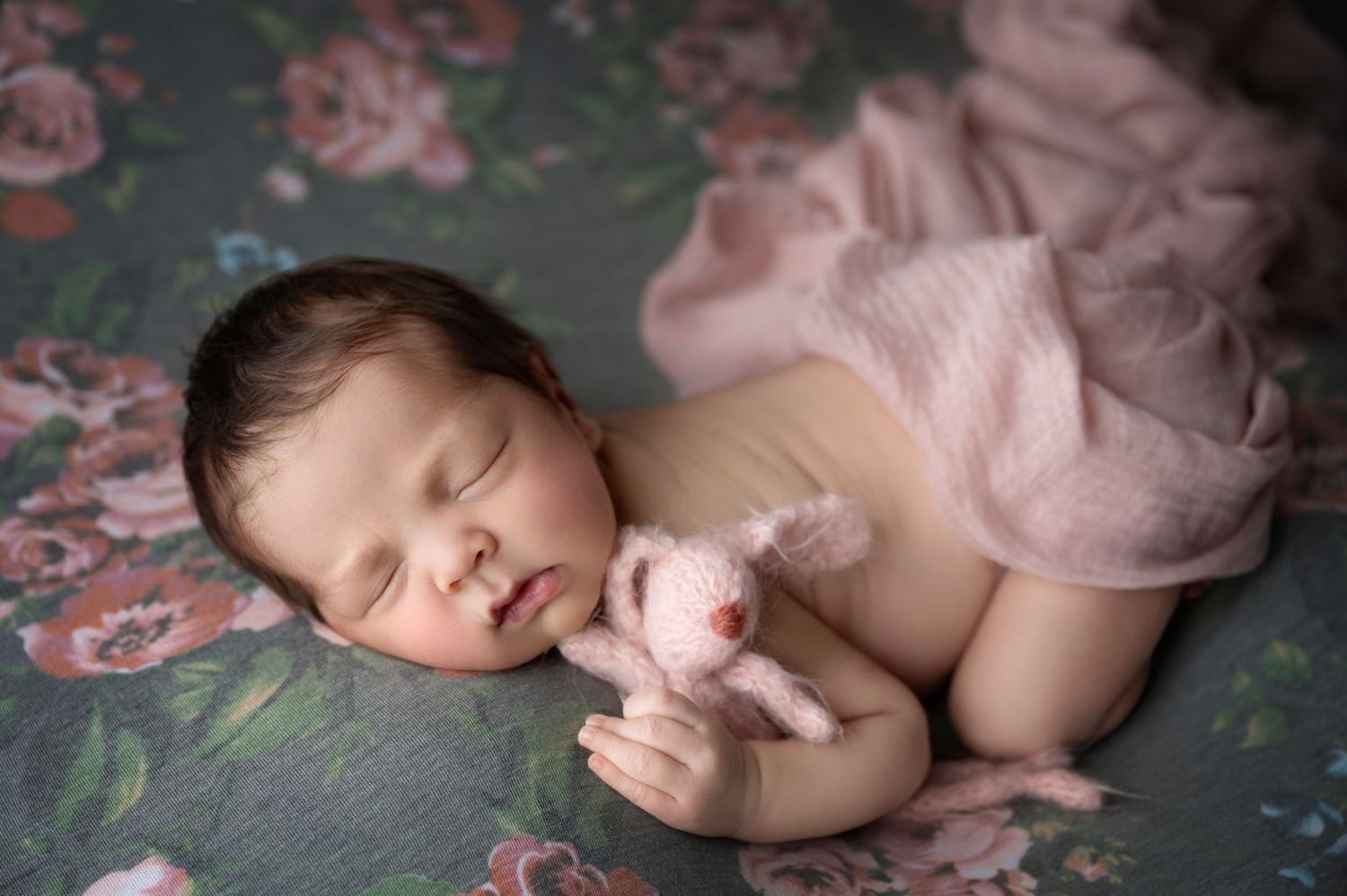 Sleeping newborn baby holding a pink stuffed bunny on a floral backdrop with soft pink wrap.