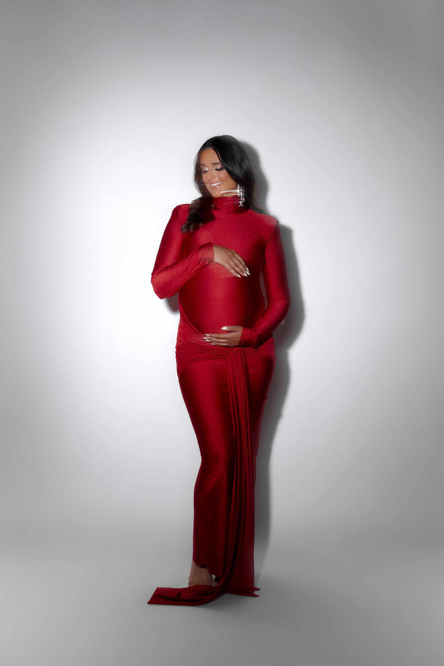 Pregnant woman in elegant red long-sleeve gown cradling her baby bump, posing gracefully against a white studio background.