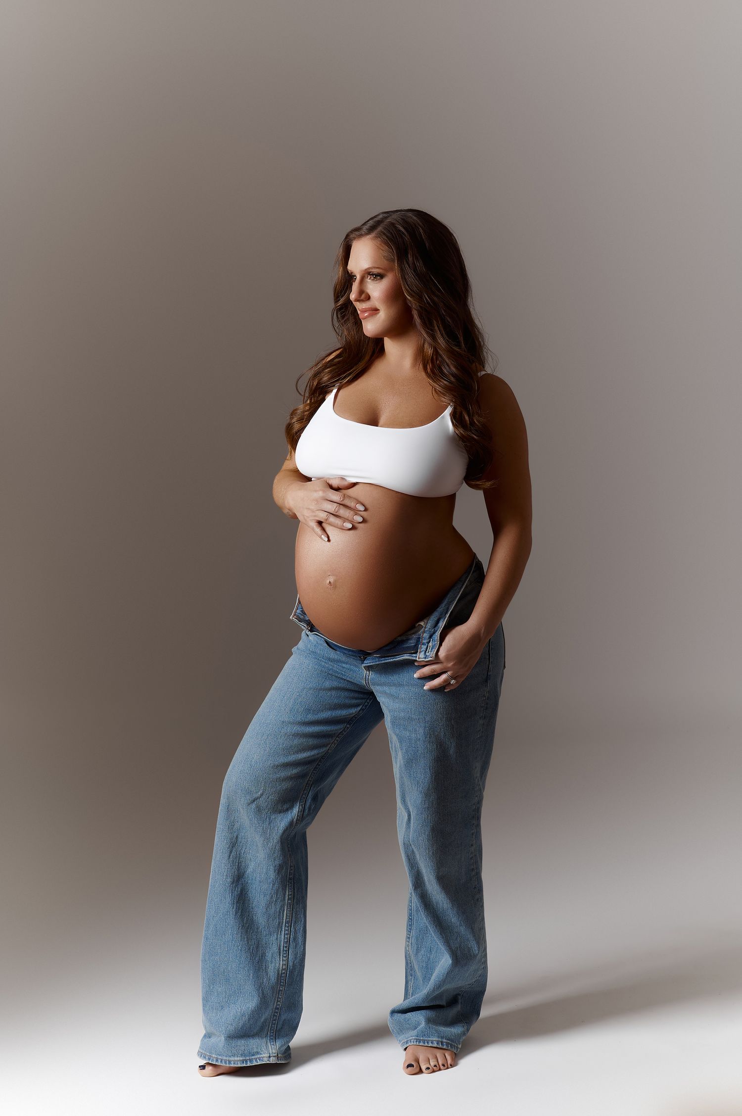 Pregnant woman in white crop top and unbuttoned jeans cradling baby bump in studio maternity photo.