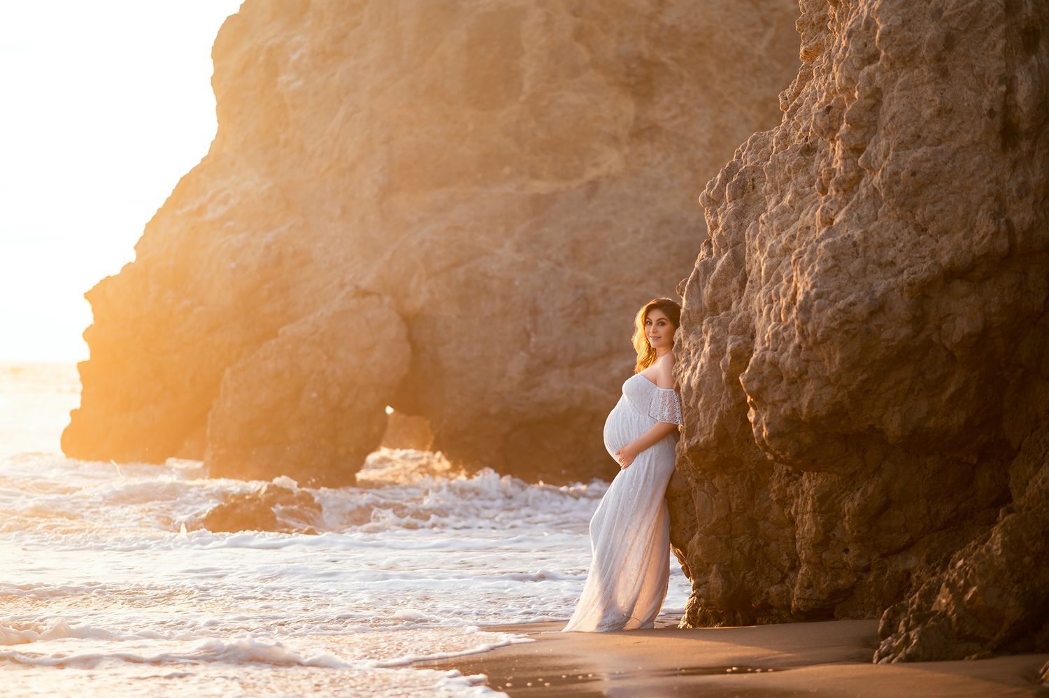 Pregnant woman in flowing white gown poses against coastal cliffs at warm golden sunset.