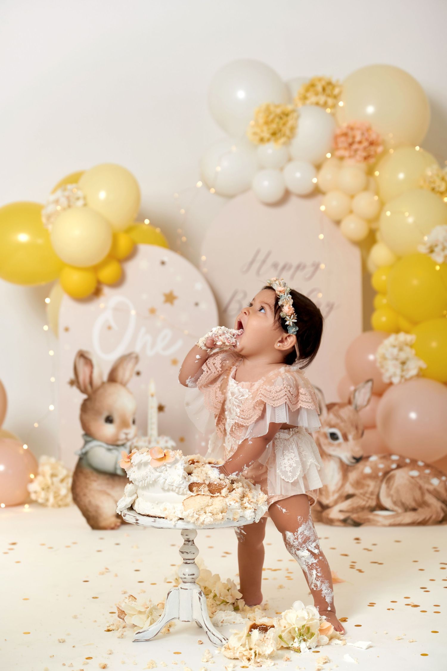 Baby girl in lace dress smashes first birthday cake surrounded by balloon arch and woodland animal decorations.