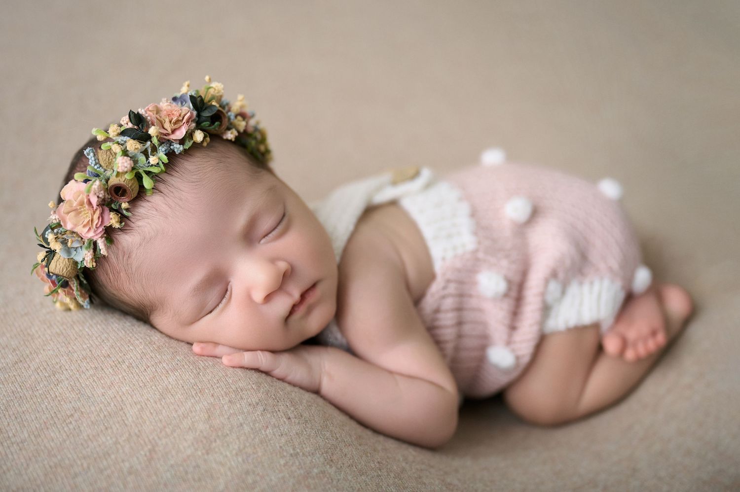 Sleeping newborn baby wearing a floral crown headband and pink knit bloomers on a beige backdrop.