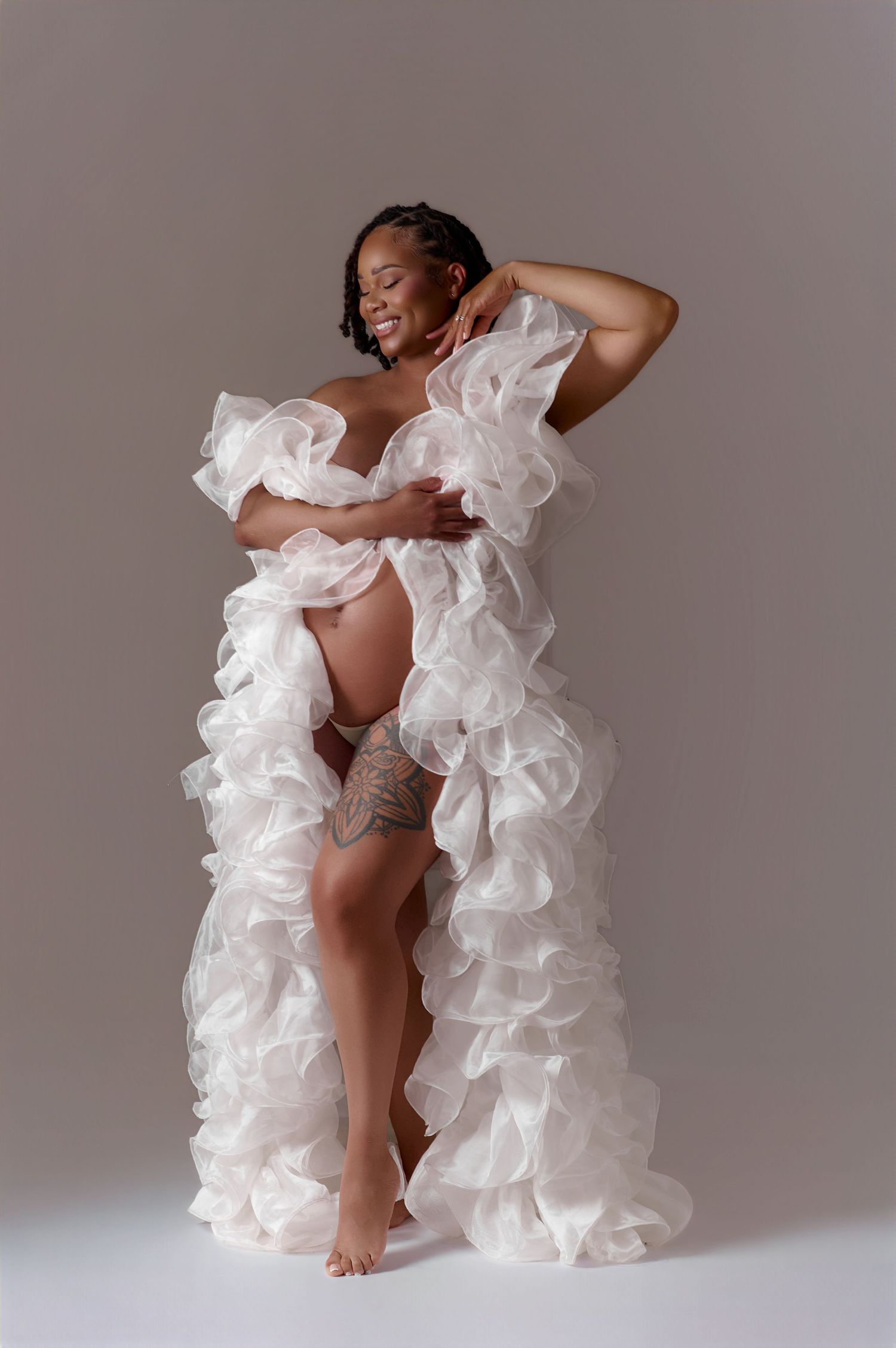 Pregnant woman in white ruffled maternity gown posing gracefully in a professional studio photo shoot.