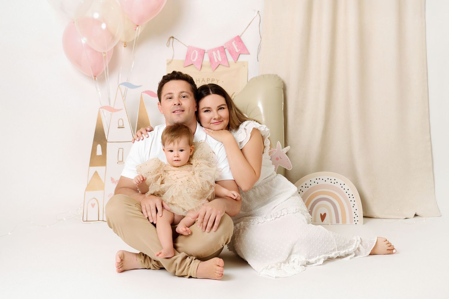 Family of three posing for a studio portrait with pink balloons and pastel decorations.