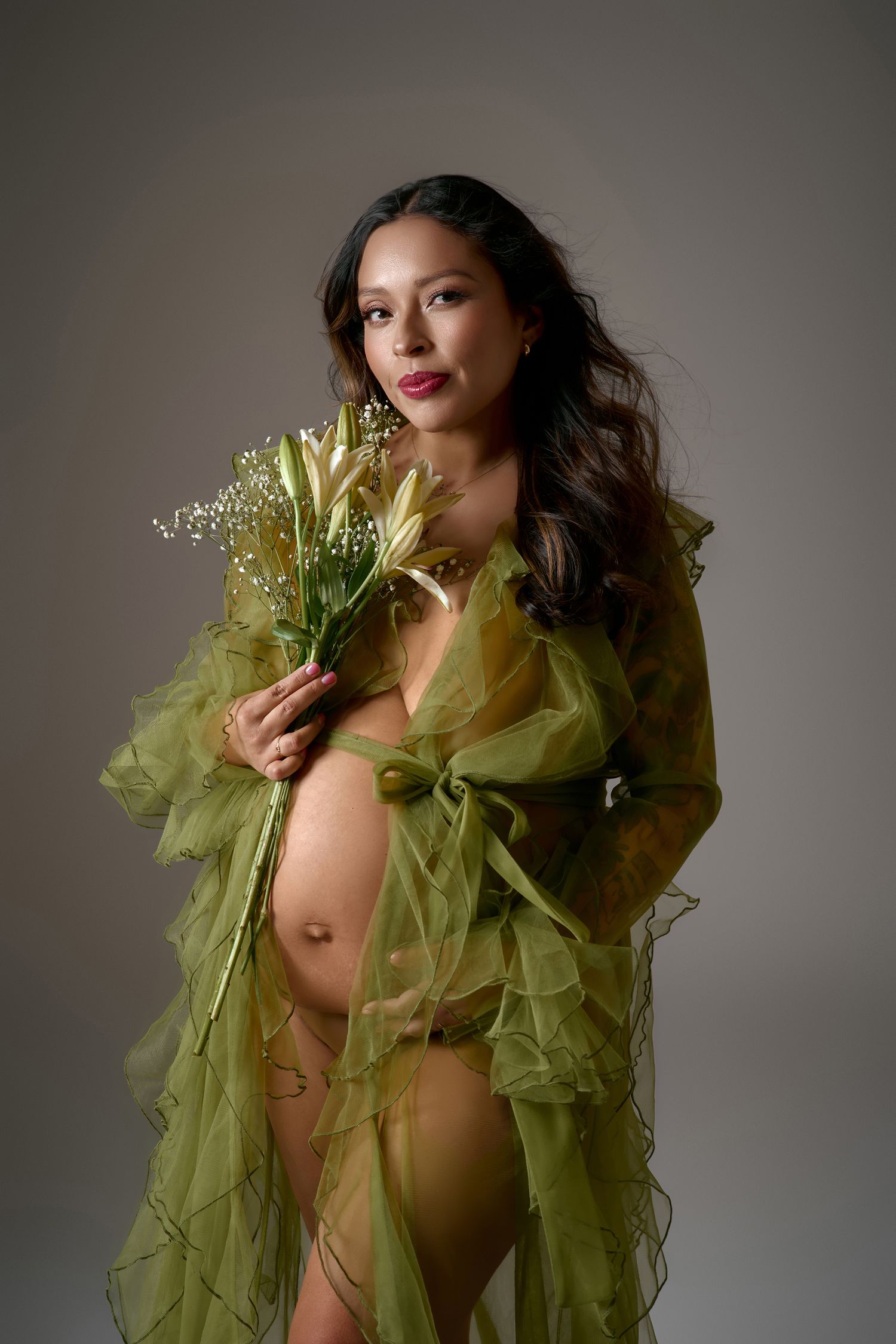 Pregnant woman in sheer olive green robe holding white lilies, posing gracefully in a studio maternity portrait.