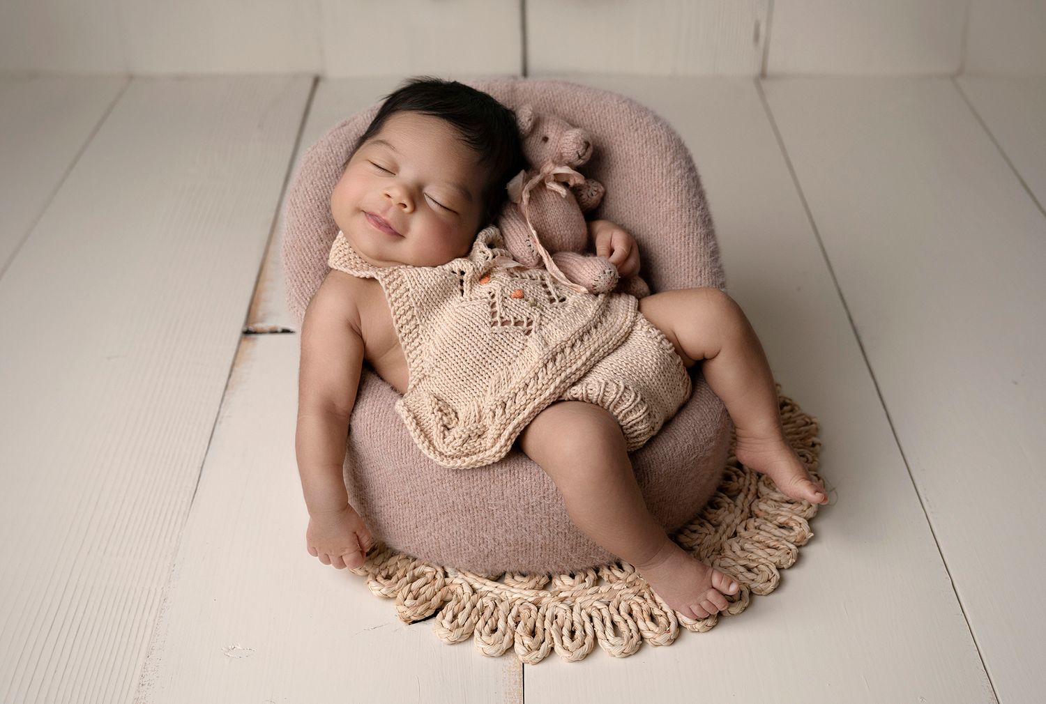 Smiling newborn baby in knit romper sleeping in round bowl prop with teddy bear on white wood floor.