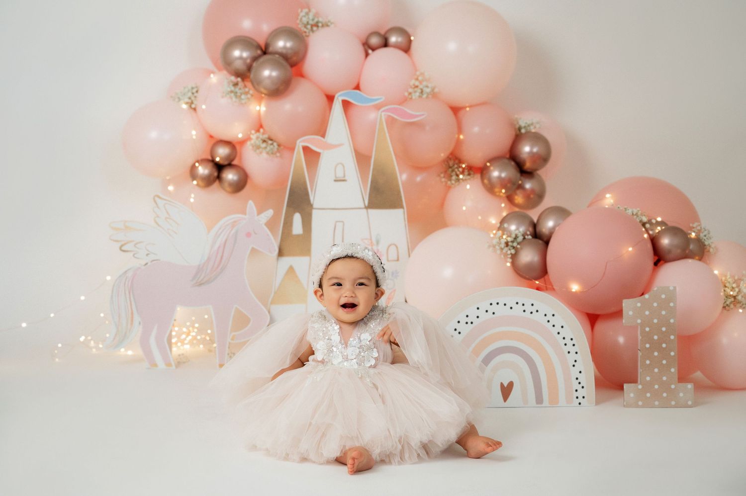 Baby girl in tutu dress sits before pink balloon arch with unicorn and castle decorations for a first birthday photo.