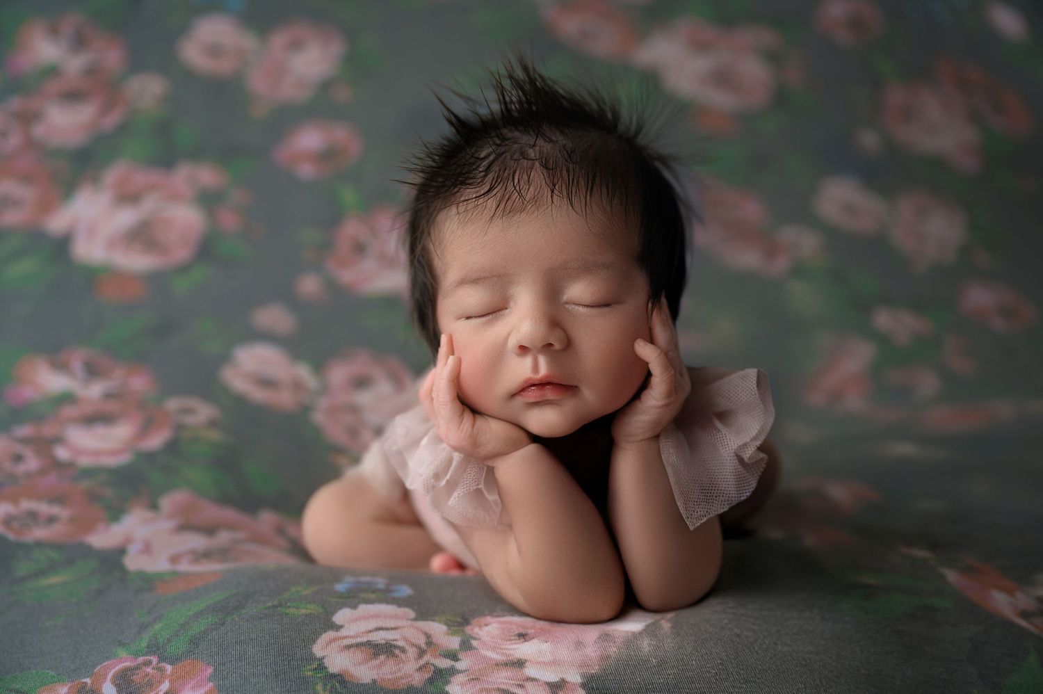 Sleeping newborn baby with dark hair posed with hands on cheeks on a floral teal backdrop.
