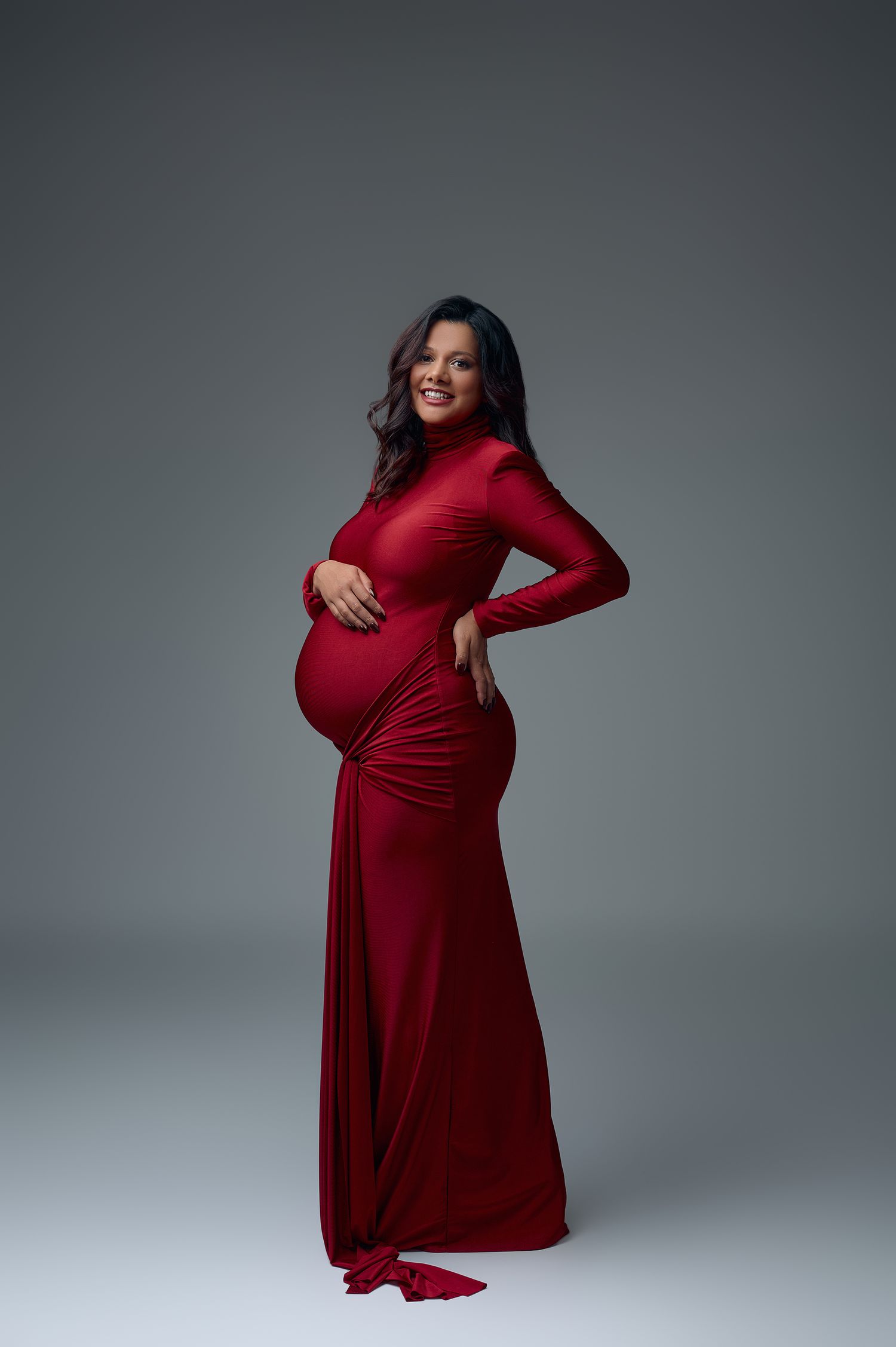 Smiling pregnant woman in elegant red long-sleeve gown posing in studio, cradling baby bump.
