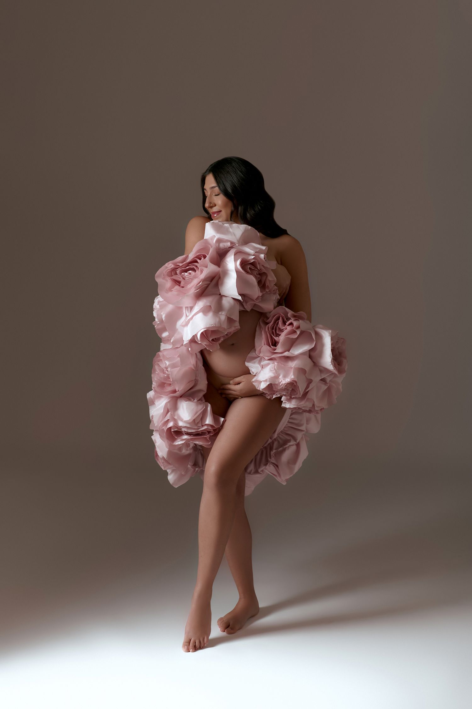Pregnant woman in voluminous pink rose petal gown posing barefoot in artistic maternity studio photo.