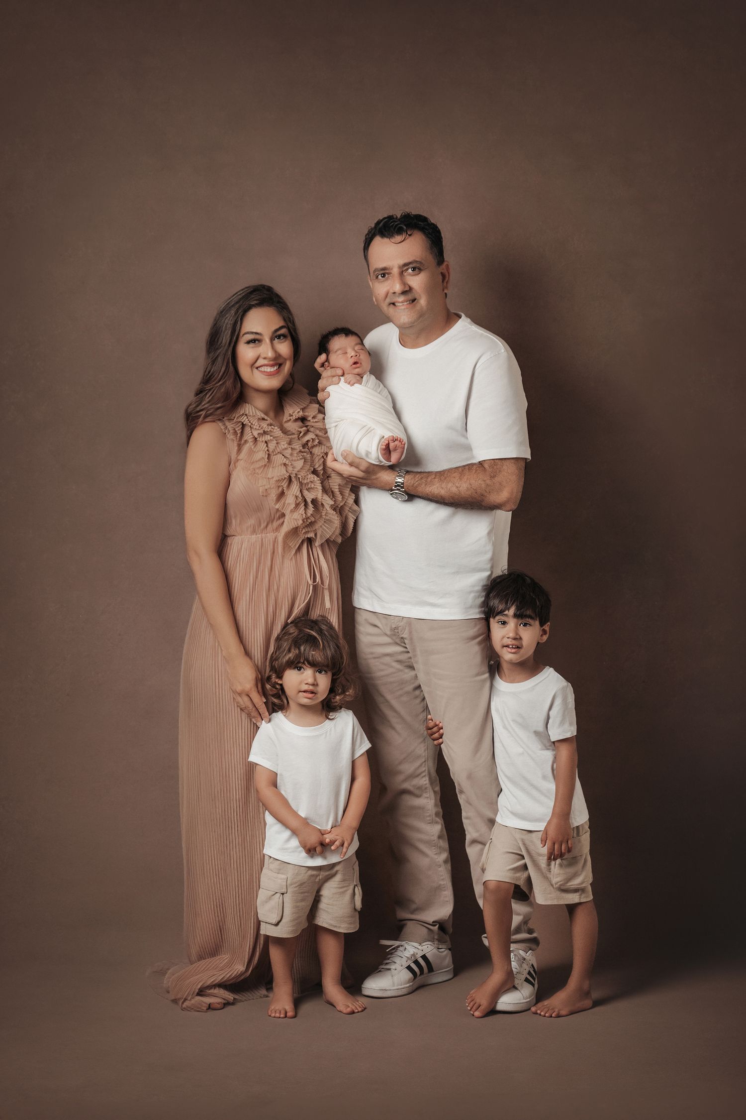Family of five posing in studio, parents holding newborn with two young boys in matching white shirts on brown background.