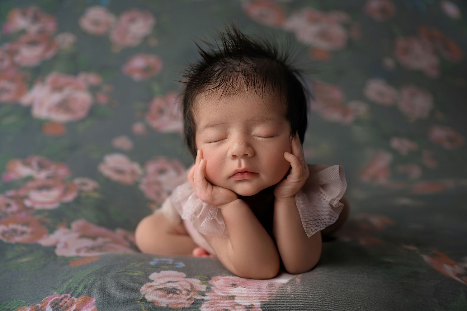 Sleeping newborn baby posed with hands on cheeks on a floral teal backdrop in a soft pink outfit.