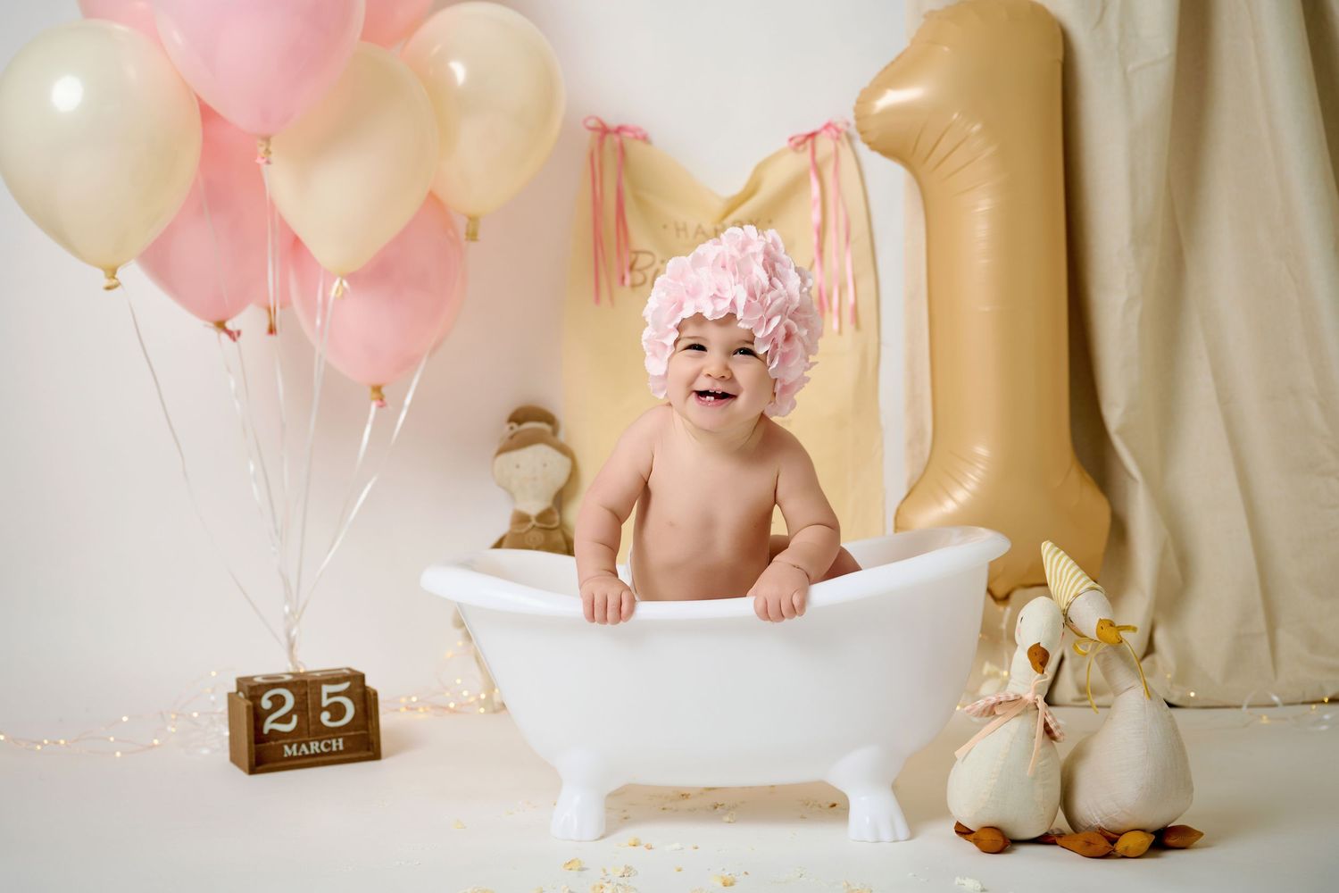 Happy baby in tiny white bathtub with pink floral hat, balloons, and number 1 decor celebrating first birthday.
