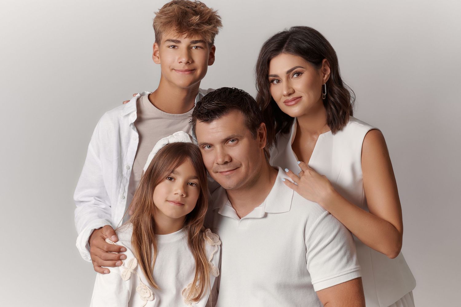 A happy family of four in white outfits posing together for a professional portrait photo against a light background.