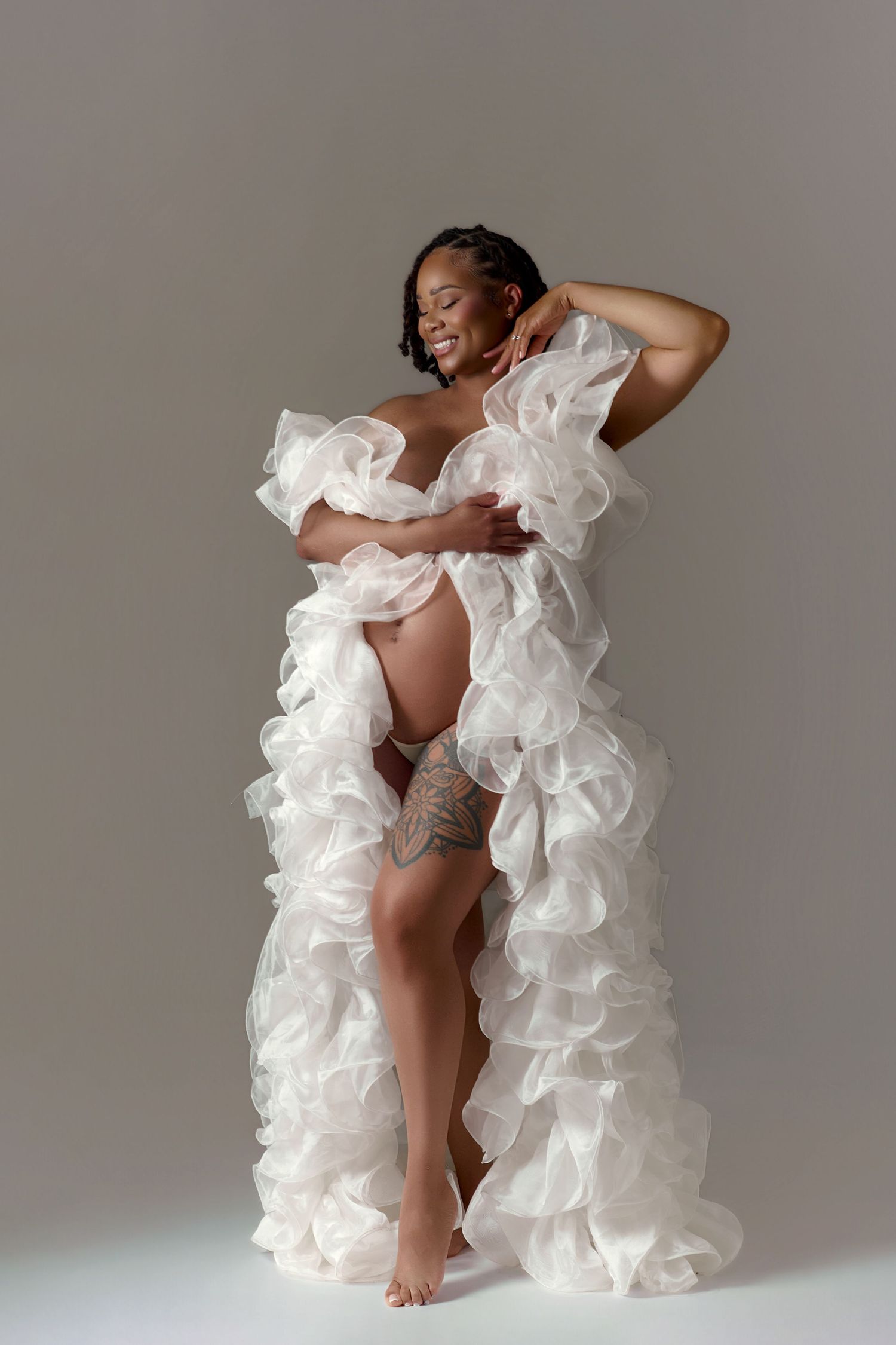 Pregnant woman in white ruffled maternity gown posing gracefully in a studio photography session.