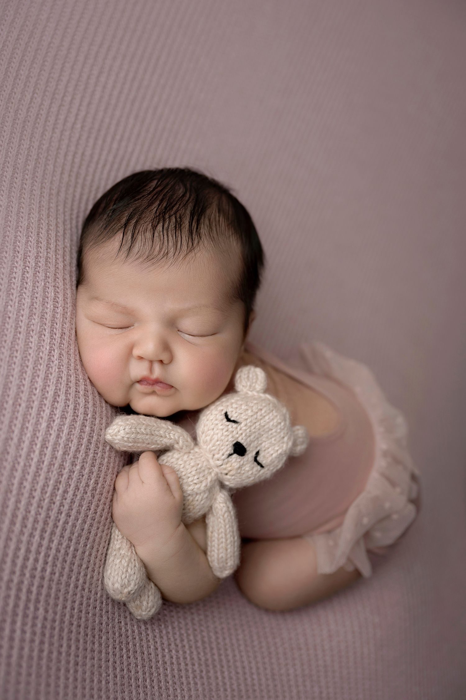 Sleeping newborn baby in pink tutu cuddling a knitted bunny toy on a soft mauve blanket.