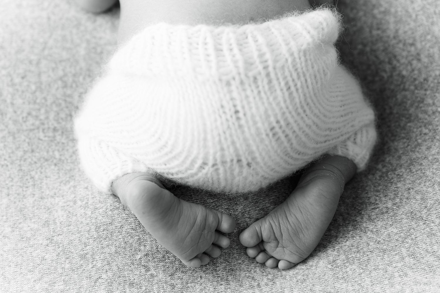 Black and white newborn baby feet and toes curled up in soft knit bloomers on a textured blanket.