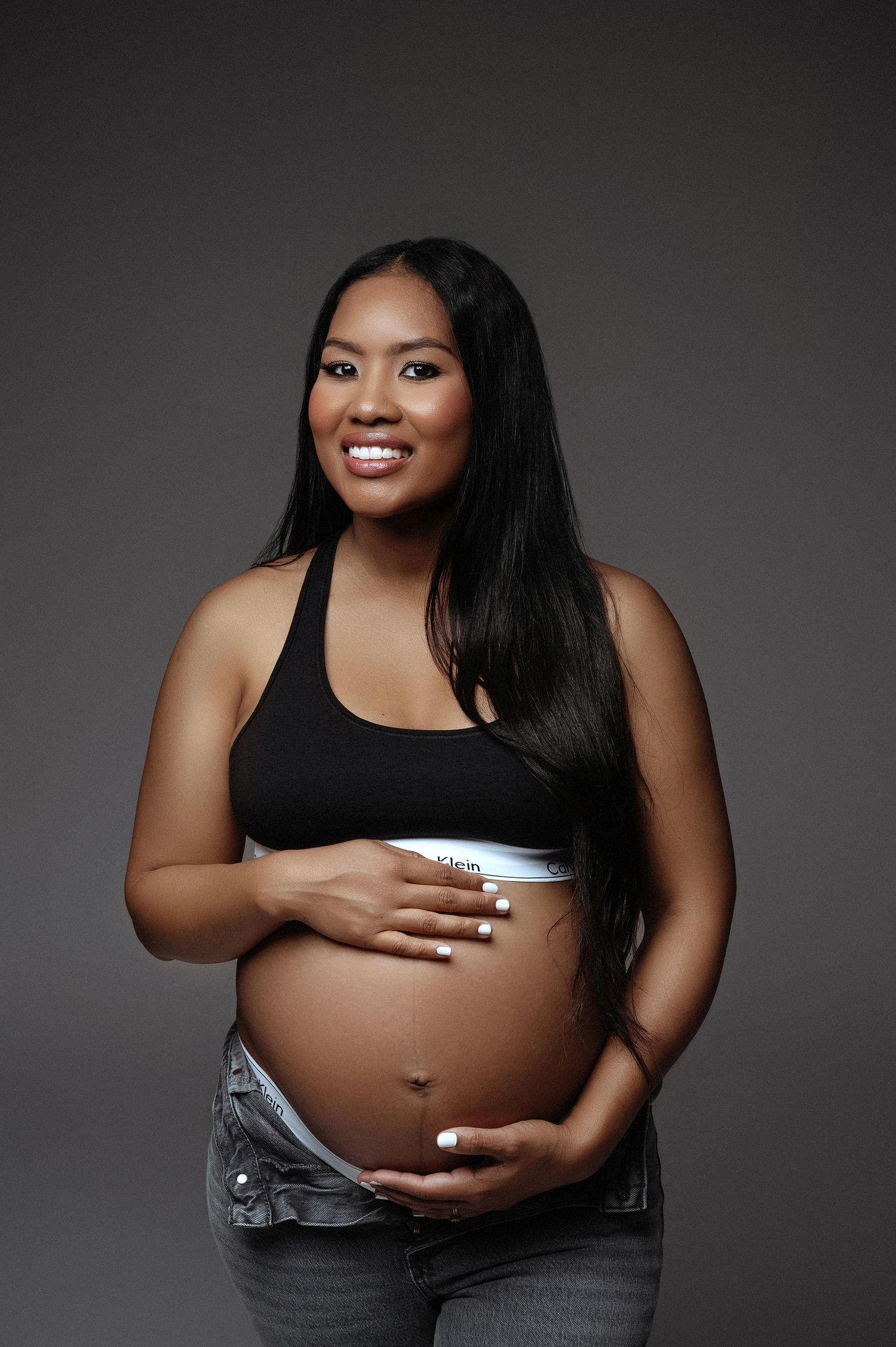 Smiling pregnant woman in black crop top and jeans cradles bare baby bump against gray backdrop.