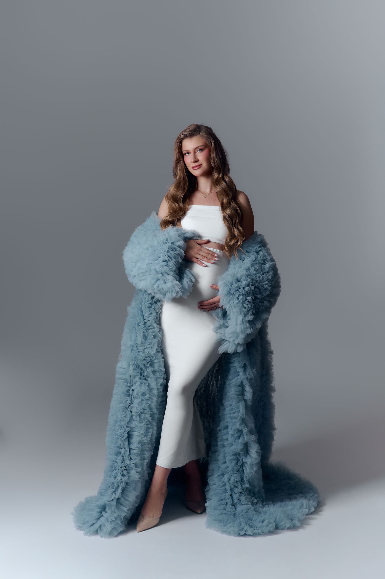 Pregnant woman in white dress wearing oversized blue fluffy coat cradles her baby bump in studio.