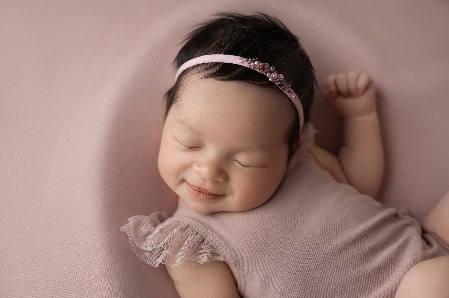 Smiling newborn baby girl in pink romper and floral headband sleeping peacefully on a soft pink blanket.