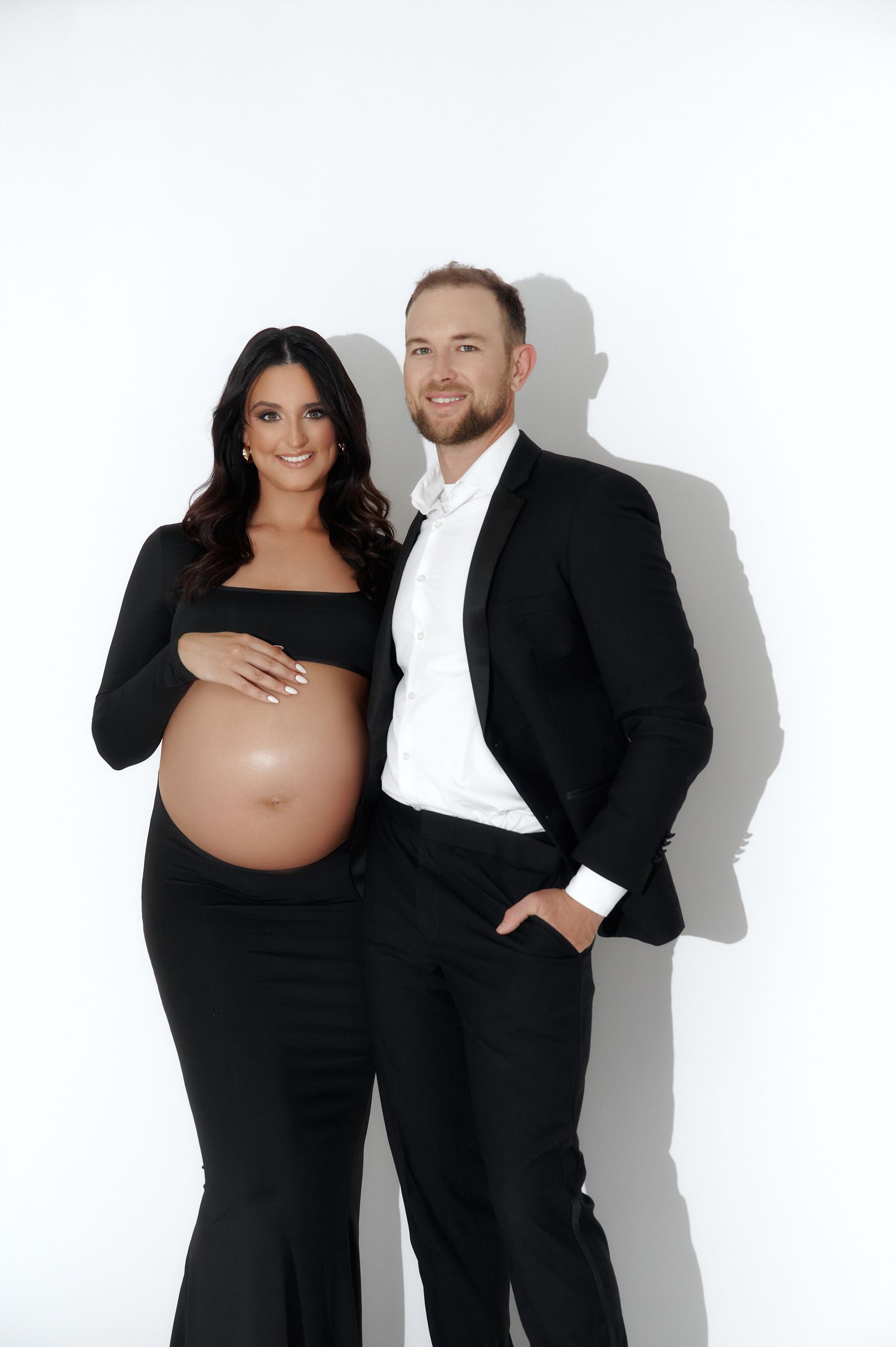 Pregnant couple in matching black outfits posing for a maternity photo shoot against a white background.