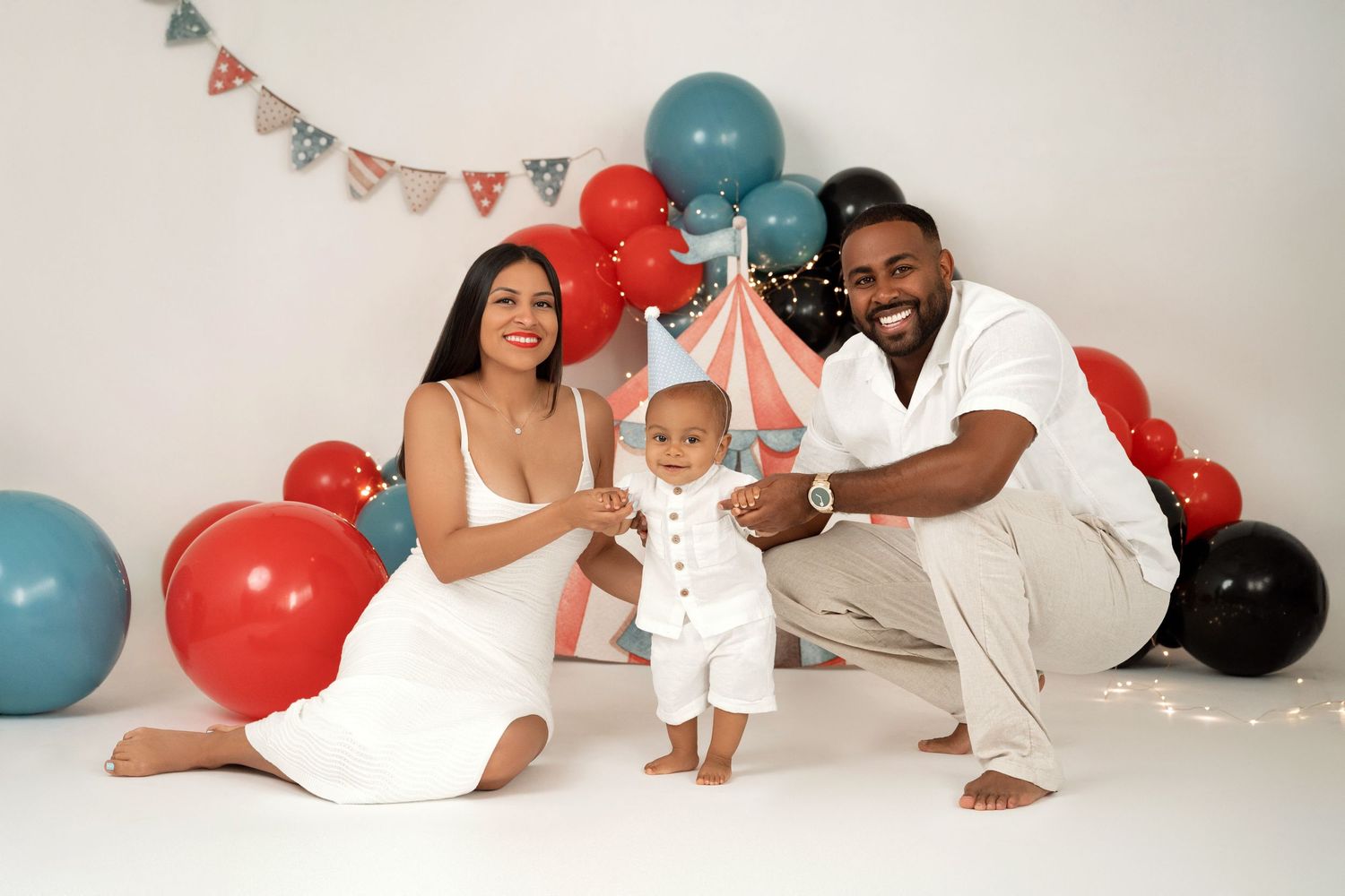 Family celebrating baby's first birthday with colorful balloons and circus tent backdrop in a white studio.