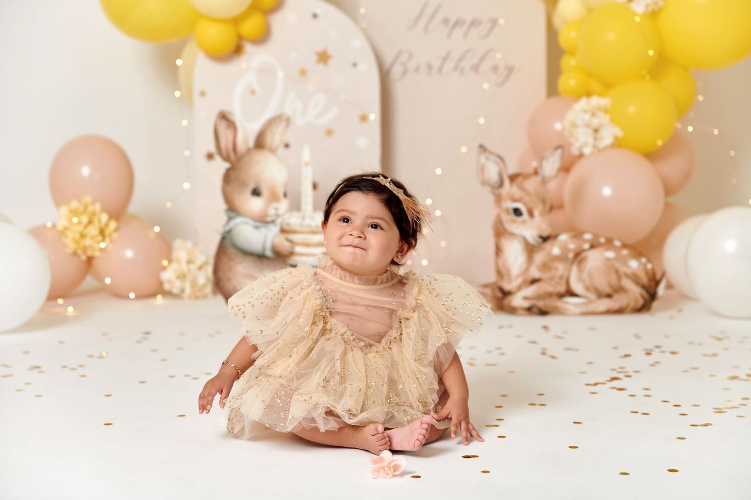 Baby girl in gold tulle dress sits smiling at first birthday cake smash with bunny deer balloons decor.