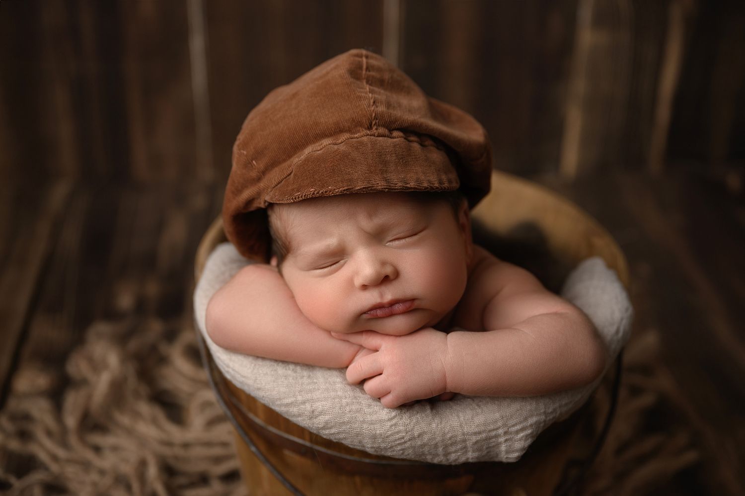 Sleeping newborn baby wearing a brown newsboy cap, nestled in a wooden bucket with a soft white blanket.