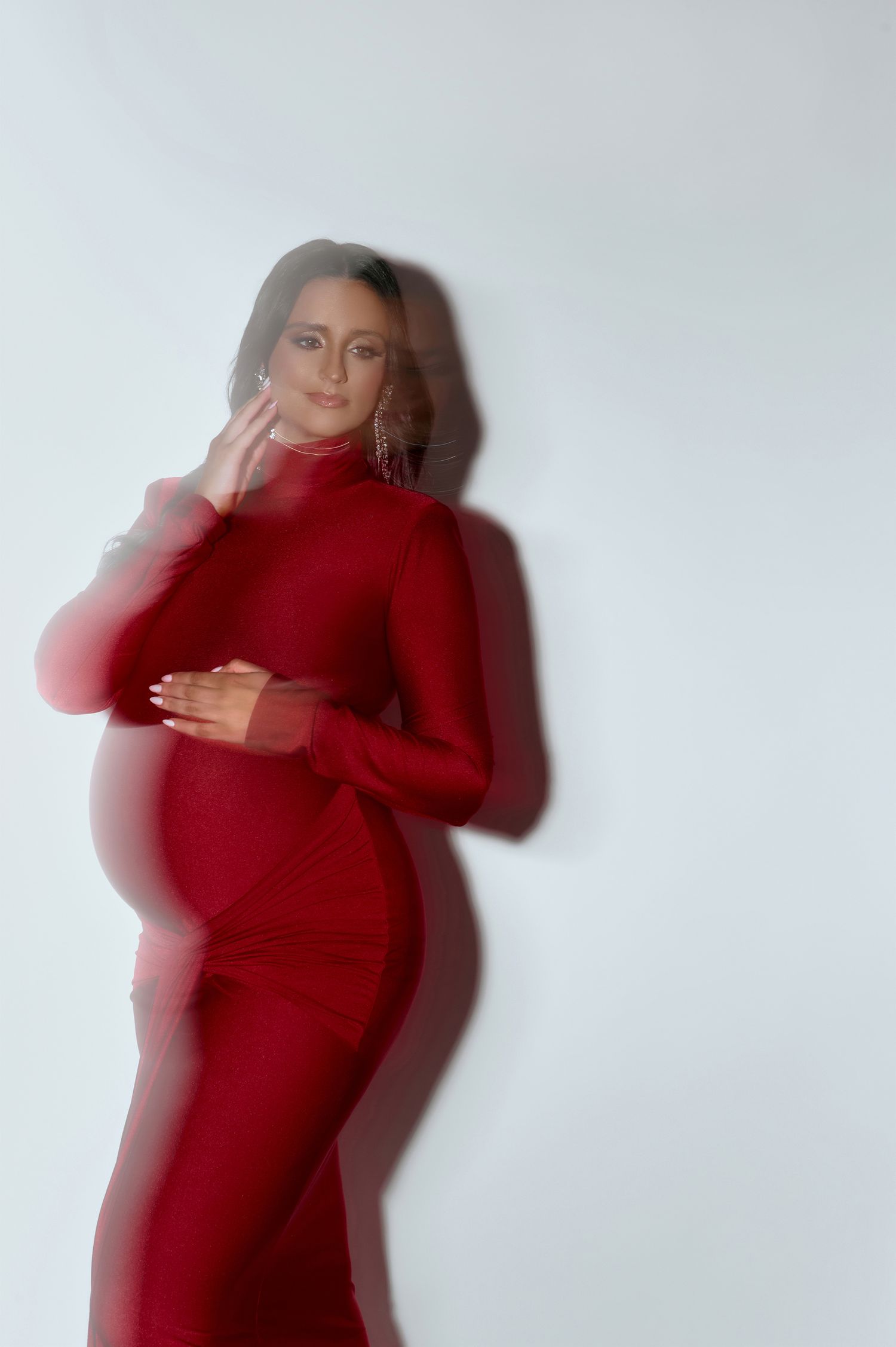 Pregnant woman in red turtleneck dress poses with motion blur effect against white studio background.