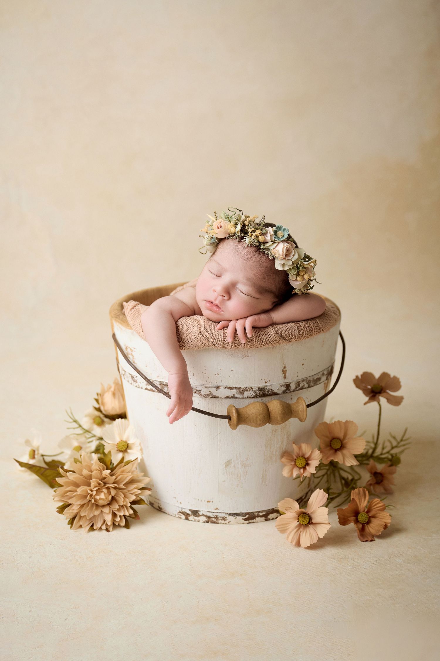 Newborn baby sleeping in a rustic white bucket with a floral crown, surrounded by soft blooms on a cream background.
