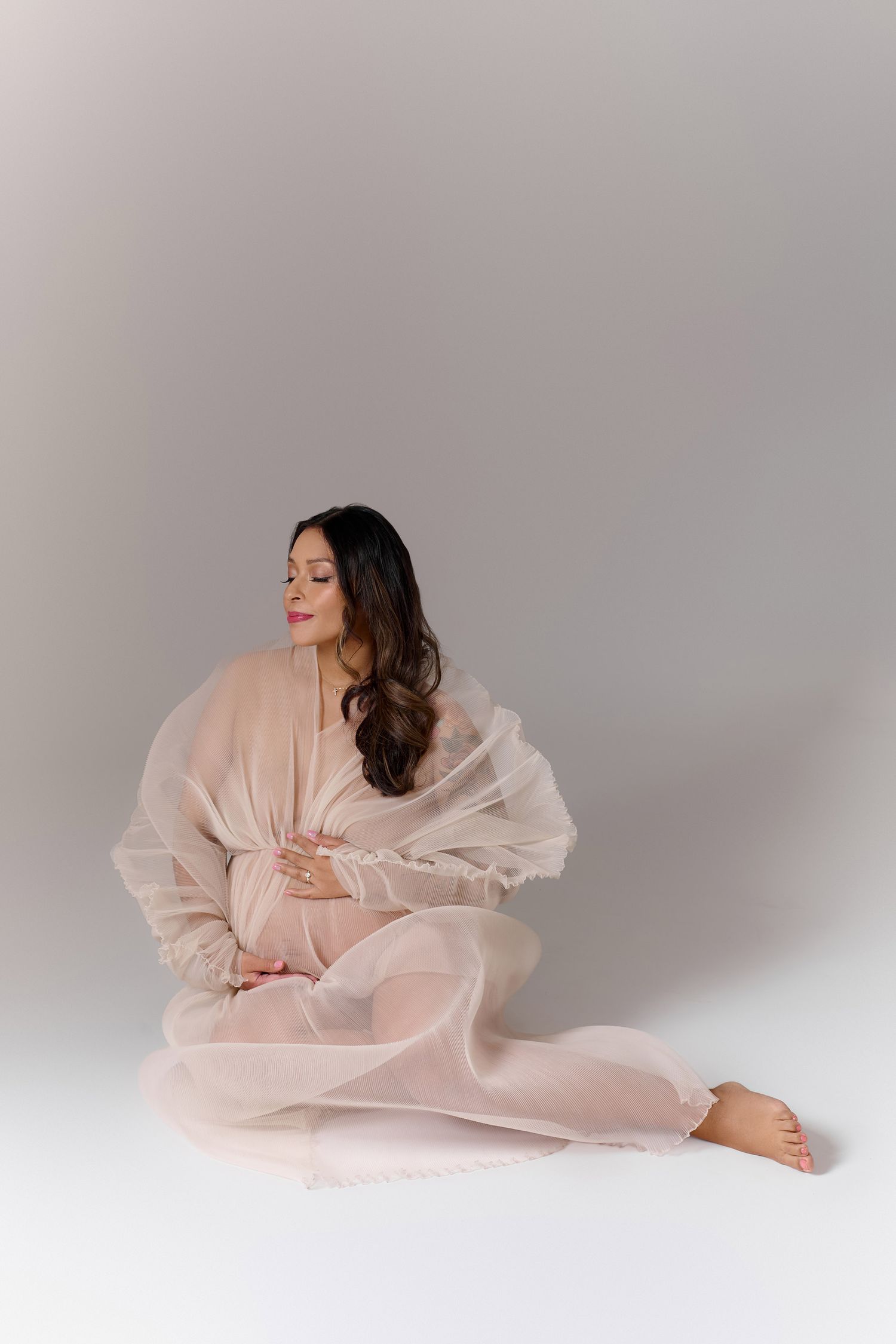 A serene pregnant woman sits on the floor wrapped in flowing blush pink fabric during a maternity photo session.