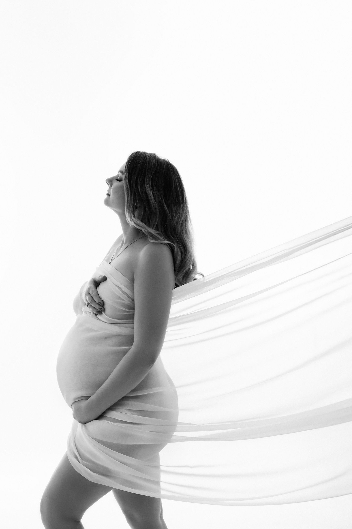 Black and white maternity portrait of pregnant woman holding bump with flowing white fabric billowing.