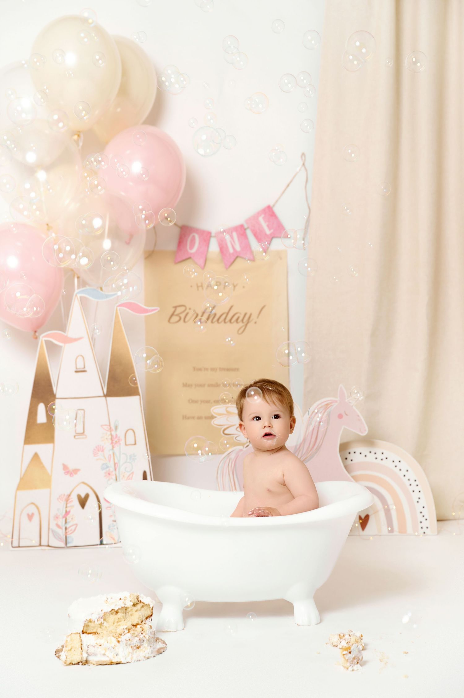 Baby girl sitting in a white bathtub at a first birthday party with pink balloons and a Happy Birthday sign.