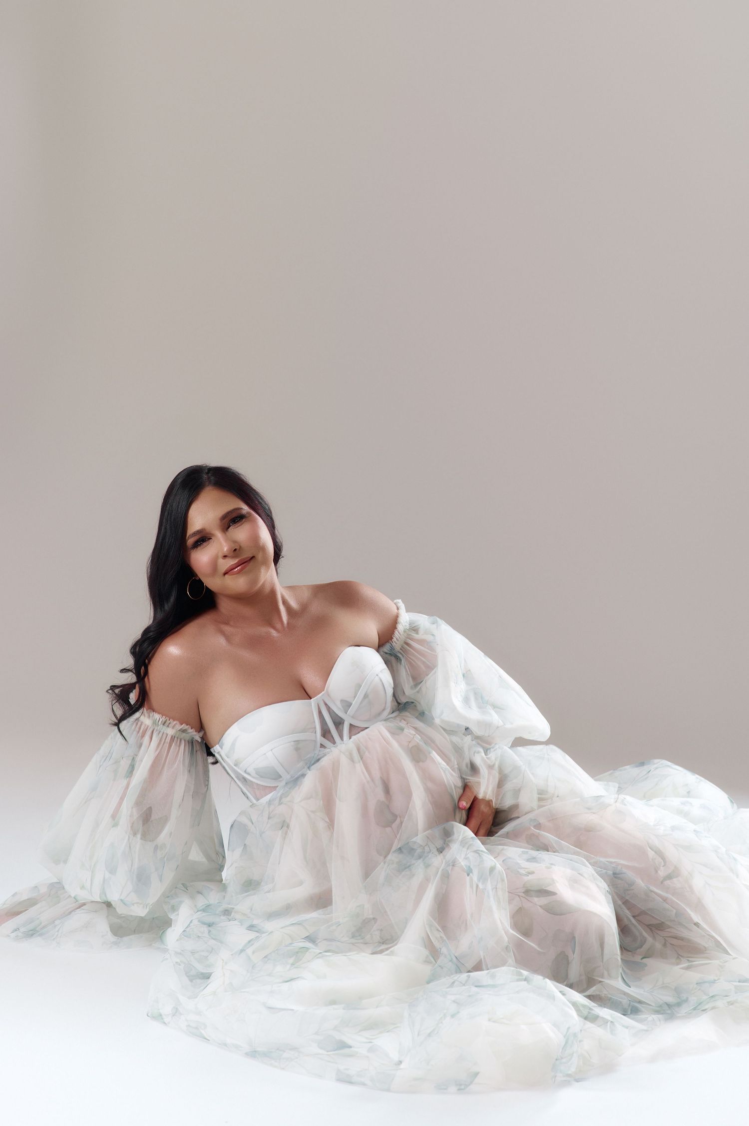 A woman in a flowing white tulle gown with floral details sits gracefully on a white studio floor.