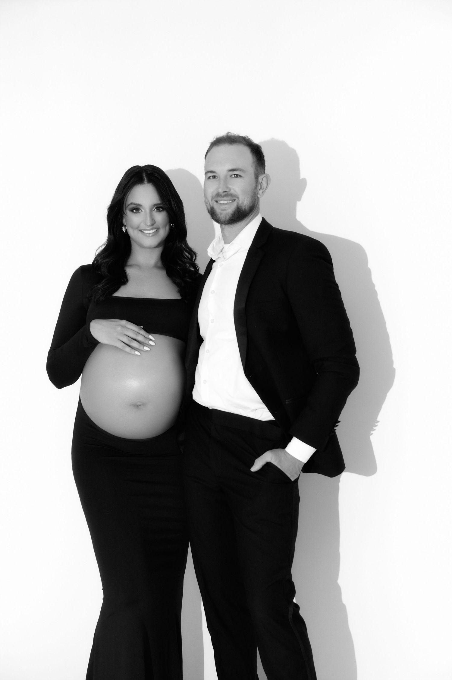 Black and white maternity photo of a pregnant woman in a black dress posing with a man in a suit against a white background.