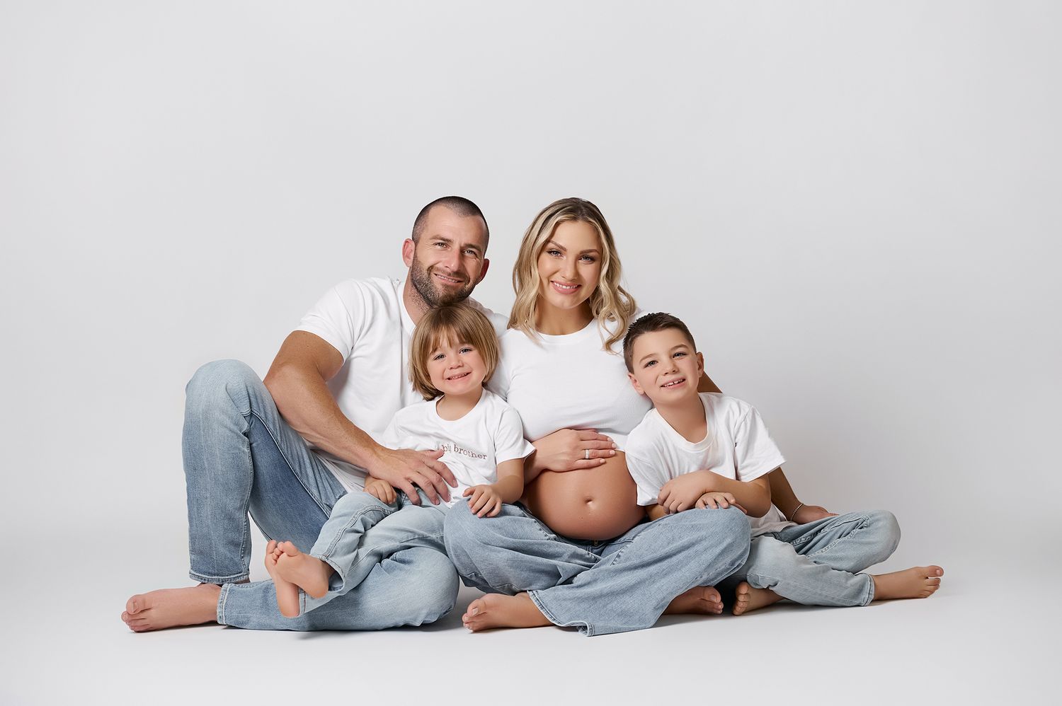 Pregnant woman in white top and jeans sits with husband and two young sons for a professional family maternity photo.